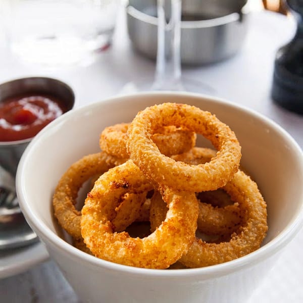 A white bowl filled with golden-brown onion rings, with a small metal cup of ketchup beside it.